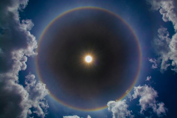The sun, haloed by a rainbow, hangs in a blue sky with clouds.
