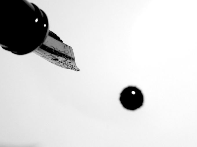 Fountain pen nib hovering over a drip of black ink on a white page